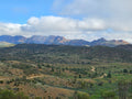 A scenic landscape photograph of rolling green and brown hills with a winding dirt road, backed by rugged mountains under a blue sky with large white clouds.