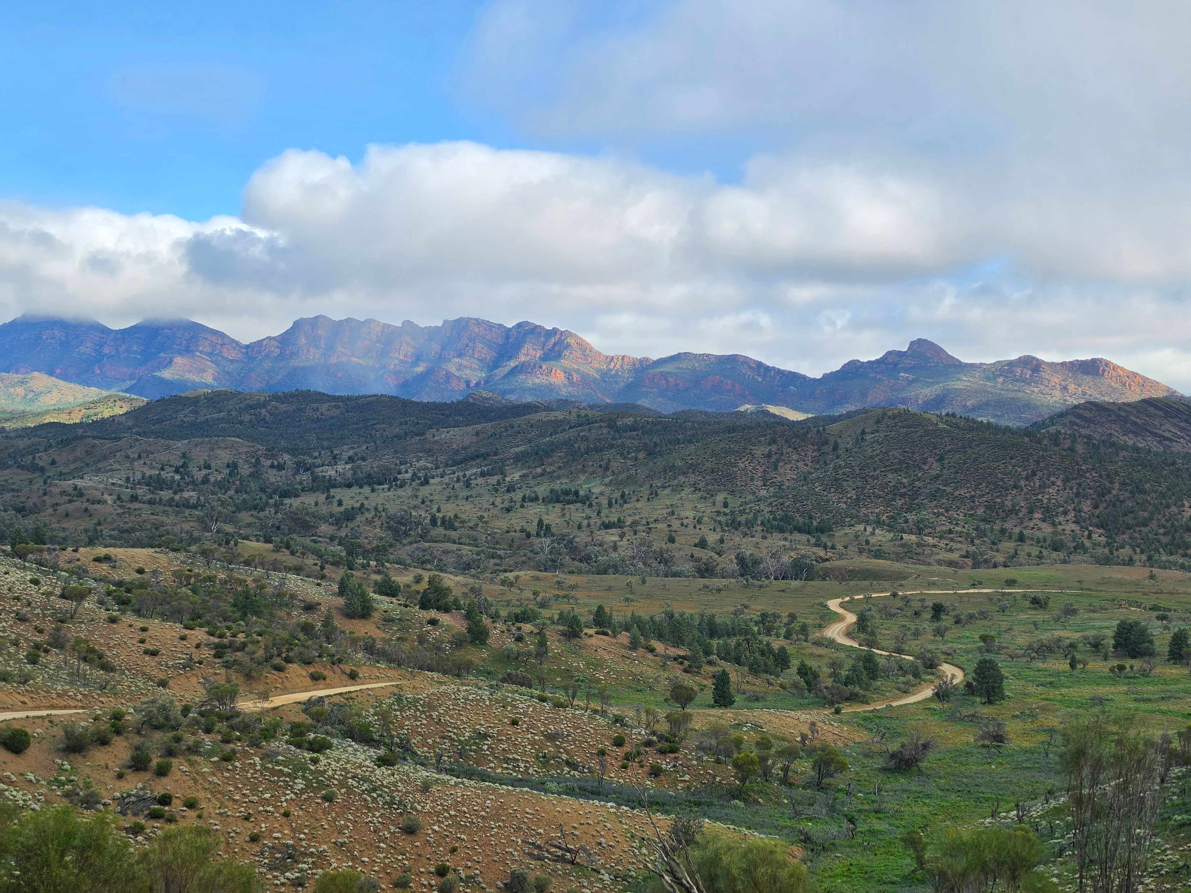 A scenic landscape photograph of rolling green and brown hills with a winding dirt road, backed by rugged mountains under a blue sky with large white clouds.