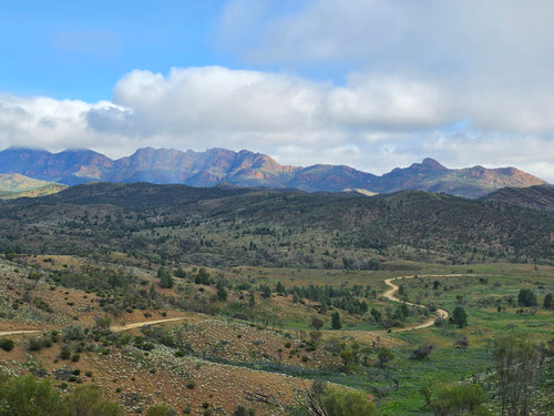 A scenic landscape photograph of rolling green and brown hills with a winding dirt road, backed by rugged mountains under a blue sky with large white clouds.